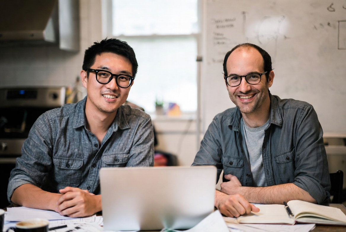 The Rootly founders working together at a kitchen table, laptop open and a whiteboard of early sketches behind them.